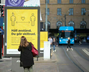 Kvinna i stadsmiljö som läser gul affisch med texten Håll avstånd. Du gör skillnad.